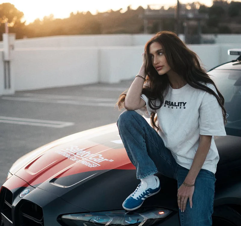 Customer wearing Reality Racing tee on a BMW safety car at sunset